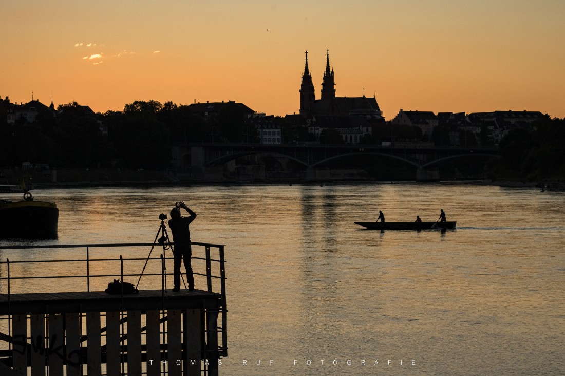 Fotograf mit Stativ am Rheinufer bei Sonnenuntergang – gelbes Licht über Basel mit Münster im Hintergrund